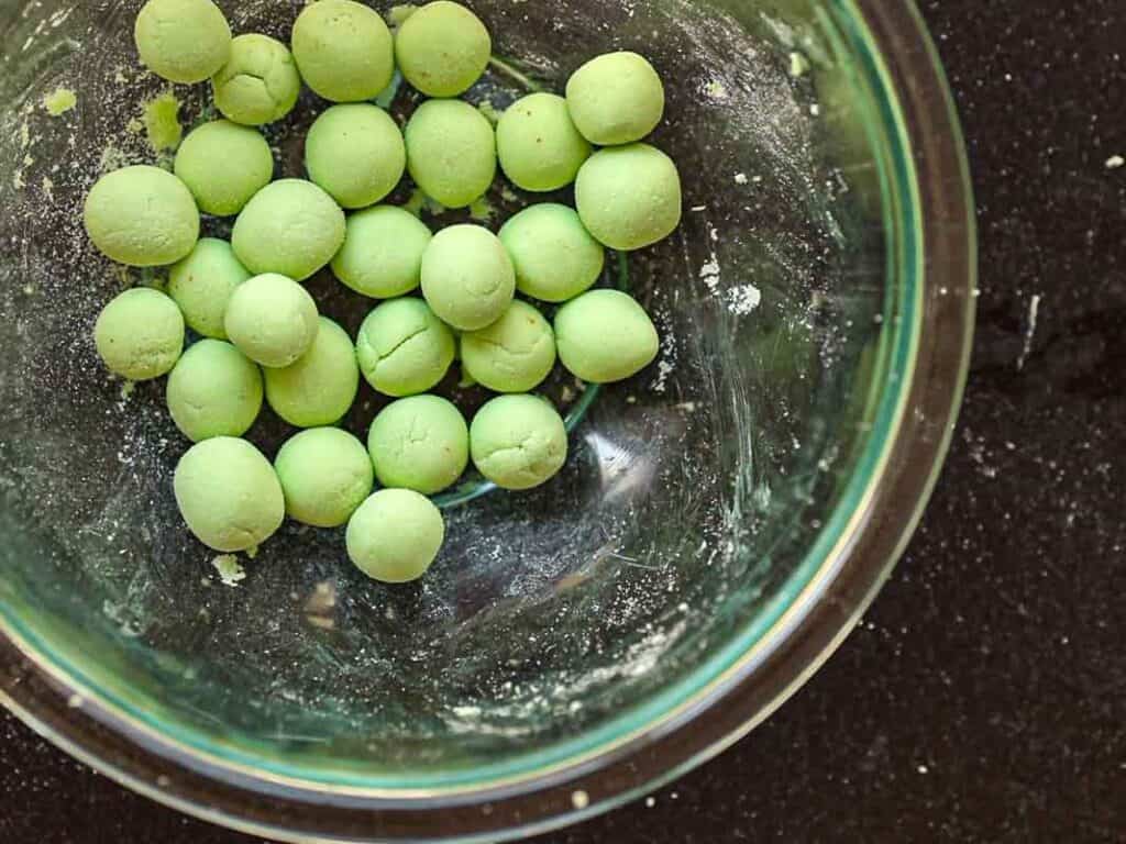 A glass bowl with small green dough balls on a dark countertop. Some flour residue is visible inside the bowl.